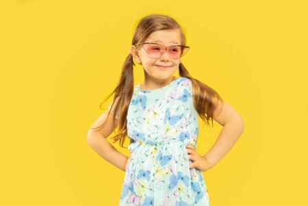 Smiling young girl in a floral dress with red sunglasses on a bright yellow background.
