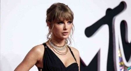 Taylor Swift posing on the red carpet at the 2023 MTV Video Music Awards wearing a fitted black asymmetrical dress and layered gold necklaces.