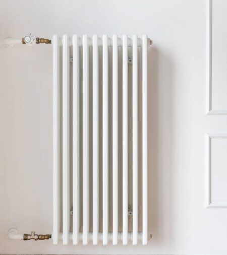 A white radiator sits on a white wall with two white pipes attached. One is attached at the top and one on the bottom.