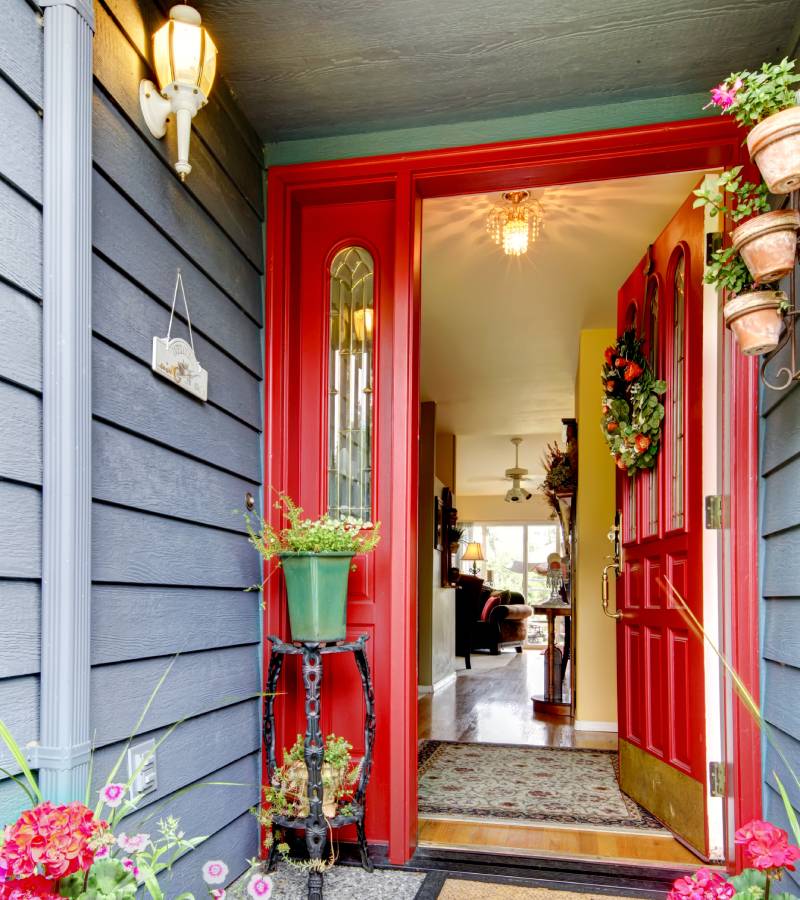 A bright-red front door is open, allowing a view into the home. There are various potted plants on the porch.