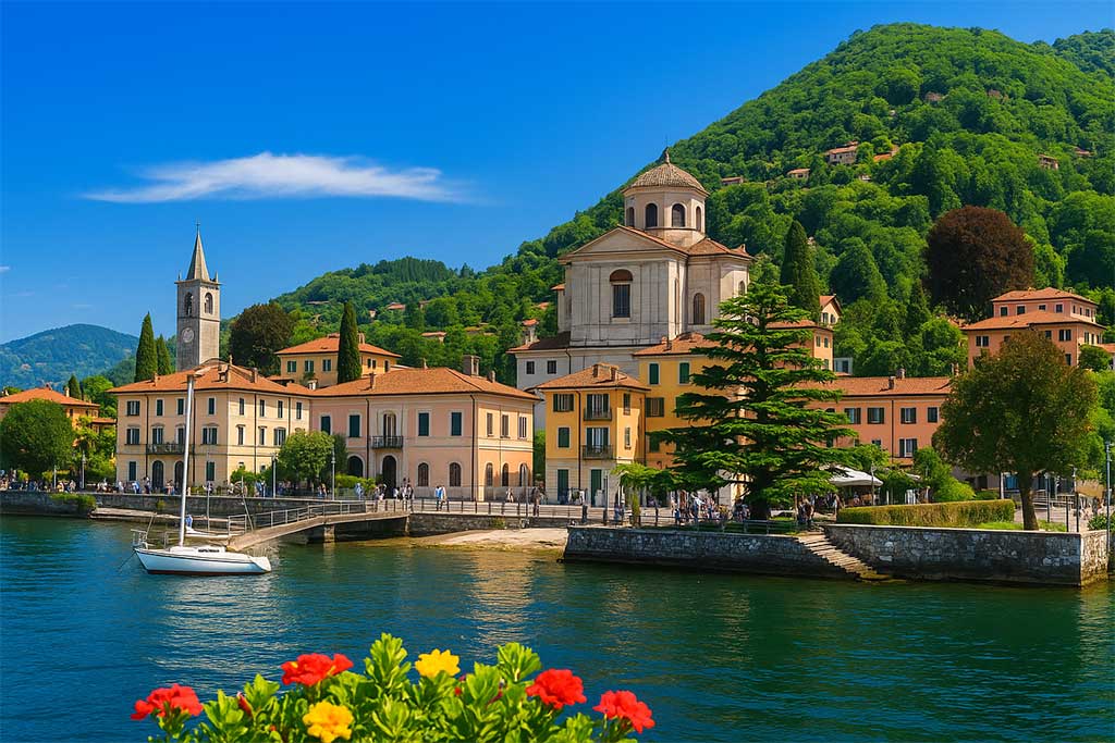 Colorful town buildings and church tower on the shores of Lake Maggiore with lush green hills behind.