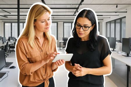 Two professional women standing in a modern office, looking at a tablet together and discussing something.