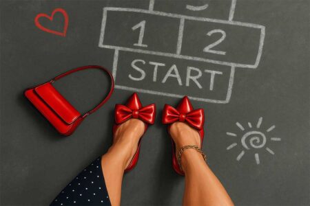A woman in red bow heels stands at the start of a chalk hopscotch grid, with a red handbag nearby on the pavement.