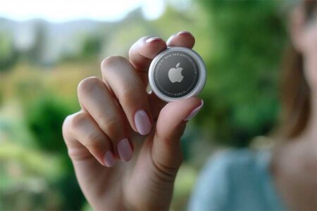 Hand holding an Apple AirTag between thumb and index finger against a soft-focus green background.