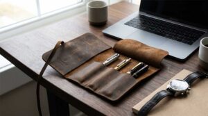 A brown leather pen roll with three pens inside sits on a wooden desk.