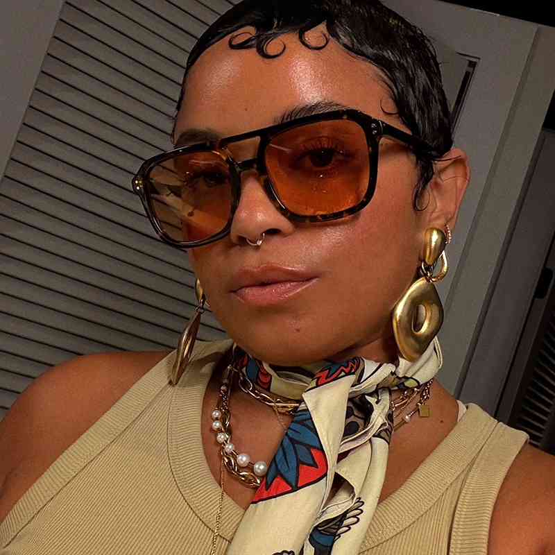 Veronica Bonilla wearing oversized amber sunglasses, gold statement earrings, and a colorful scarf